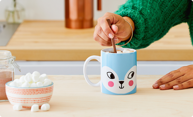 Hand stirring hot cocoa with marshmallows in a Cricut-customized mug on a kitchen counter