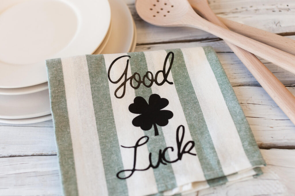 Gray and cream colored dinner napkin with Good Luck and a four leaf ironed on in black with Cricut, sitting on a neutral wood table