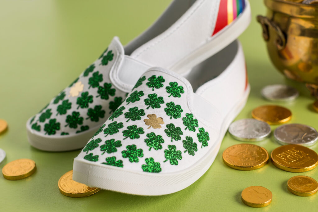 White canvas shoes decorated with green glitter four leaf clovers and one gold glitter four leaf clovers made with Cricut