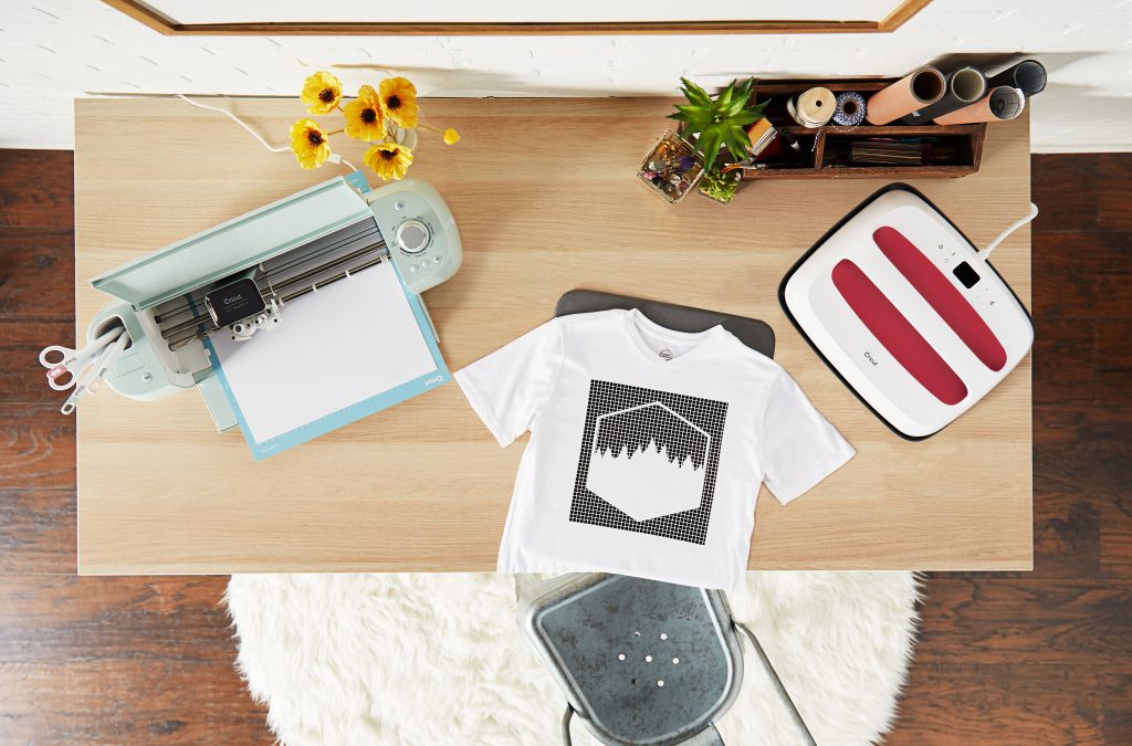 A Cricut Explore machine sits on a table with an EasyPress and a recently made customised T shirt